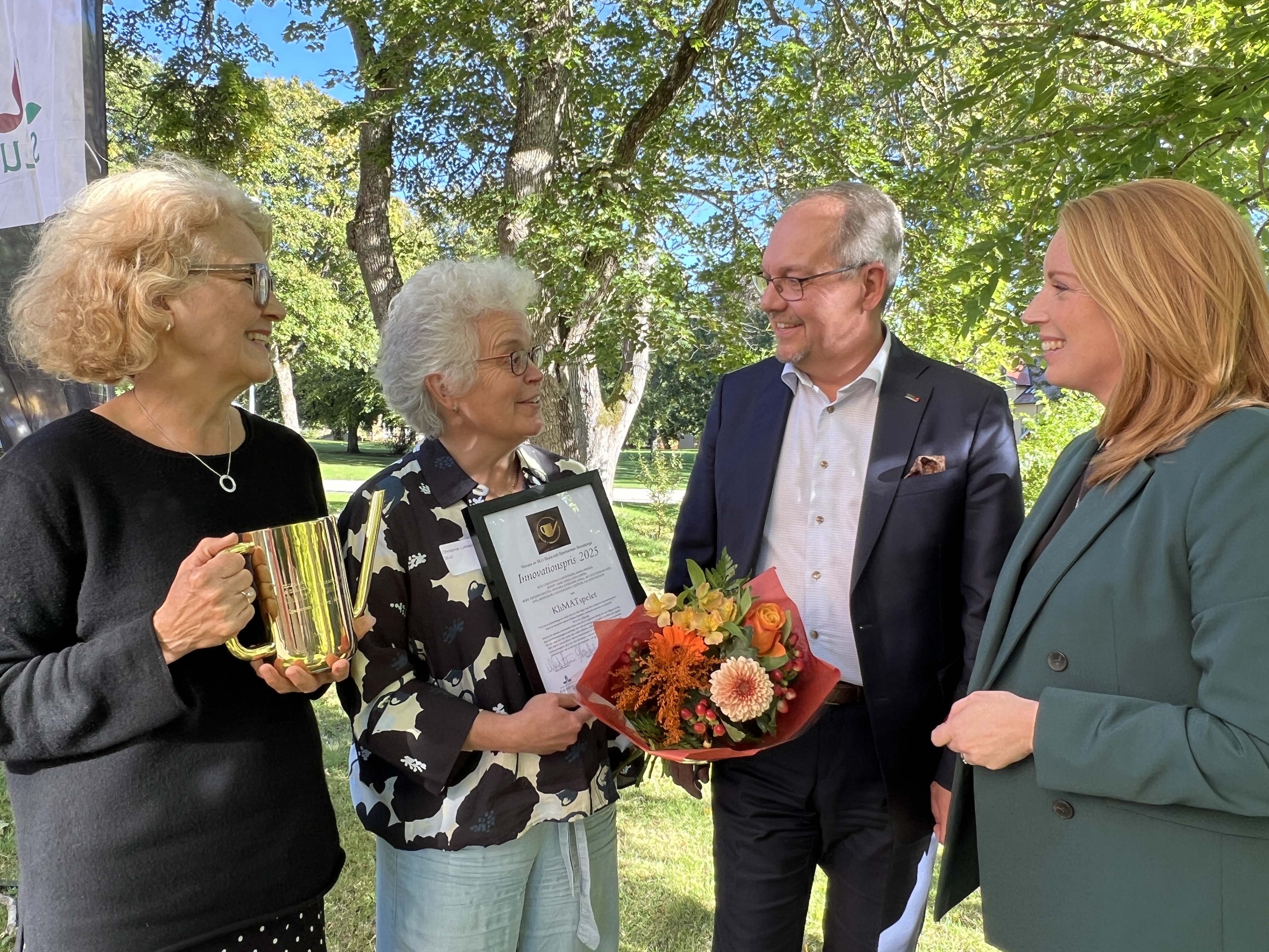 Fyra personer utomhus. En kvinna håller i en vattenkanna i mässing, en kvinna håller i ett inramat prisbevis och blommor. 