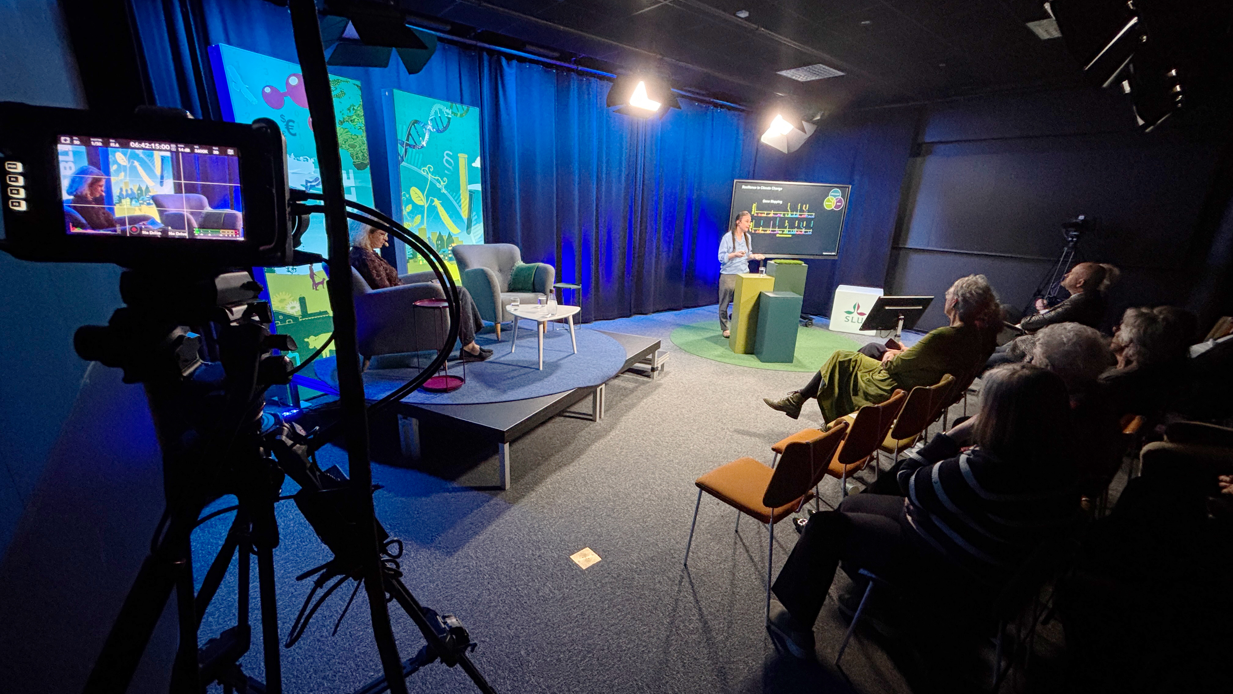 En person föreläser vid en skärm i en tv-studio, med publik och kameror.