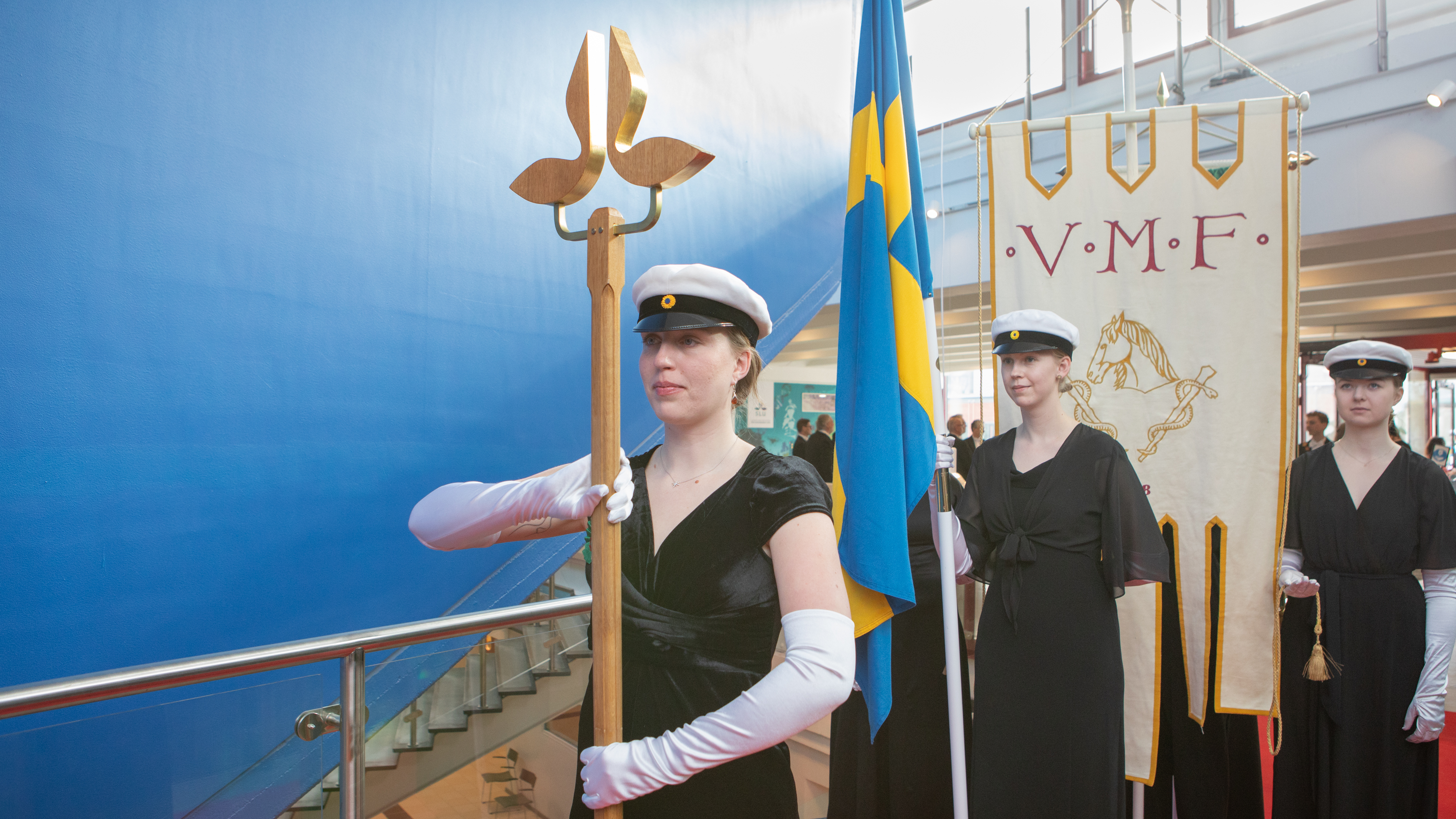 Unga högtidsklädda personer i studentmössor och långa vita handskar håller i färgglada fanor. Personen längst fram håller en trästav med ett emblem. 