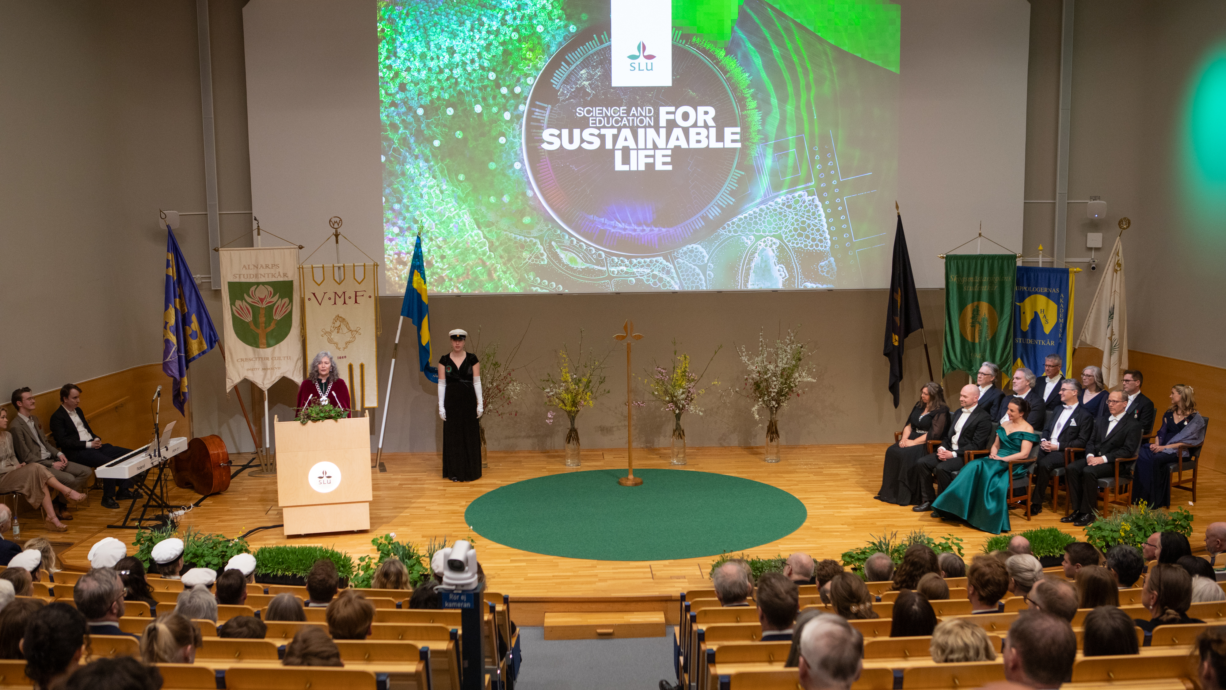 På scenen, i en talarstol står en kvinna i röd sammetskåpa och talar inför en publik. På väggen projiceras ett färggrant bildkollage. På scenens högra sida sitter 11 högtidsklädda personer i karmstolar. 