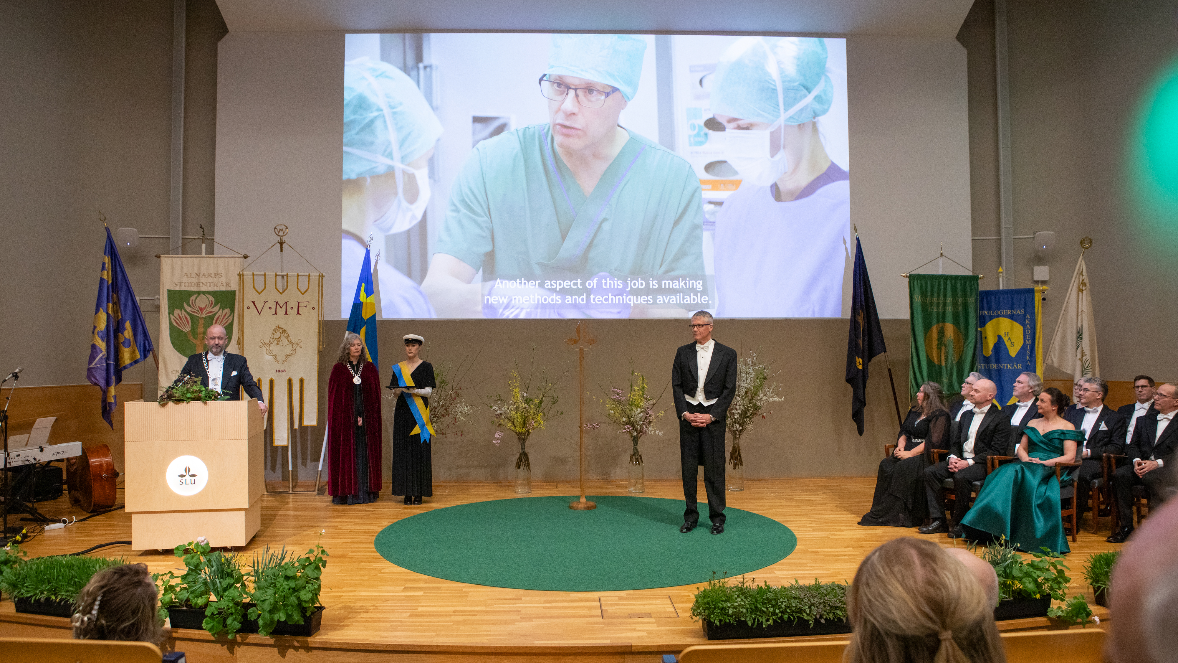 Högtidsklädda personer på en scen, framför en publik. På väggen bakom scenen projiceras en video.