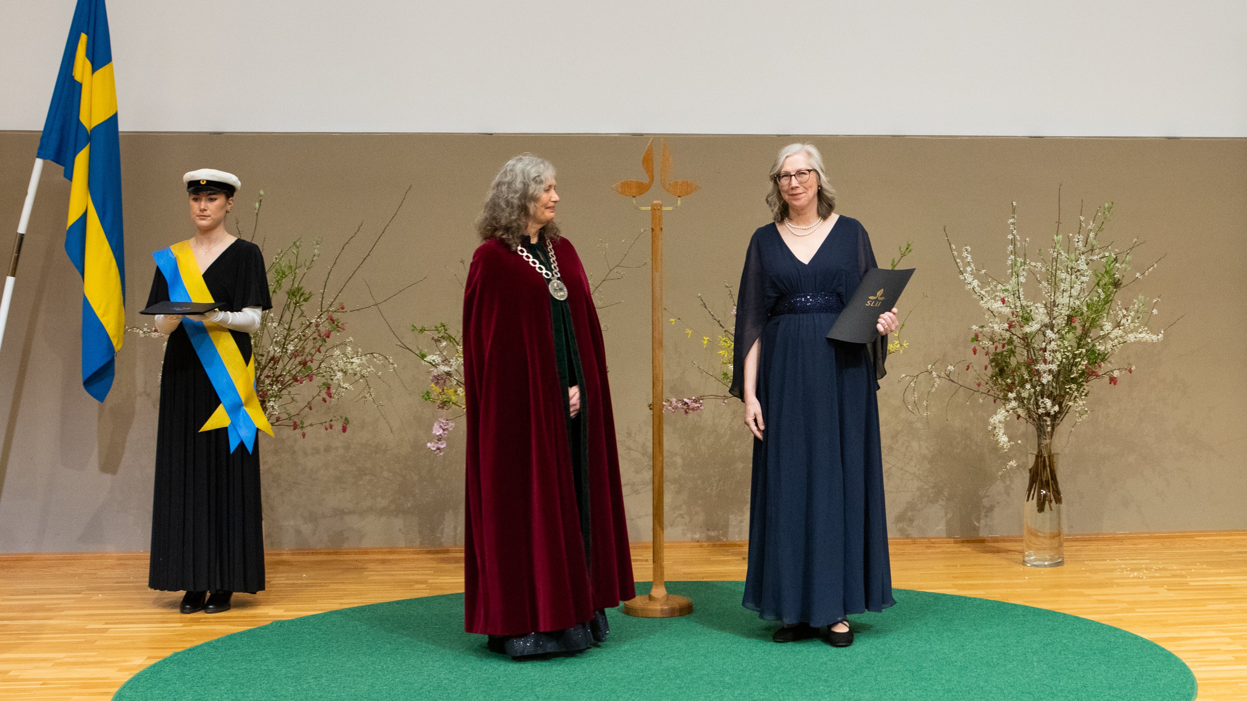 Tre högtidsklädda personer på en scen. En svartklädd i studentmössa och vita handskar, en i röd sammetskåpa och rektorskedja och en i mörkblå klänning. Den sistnämnda håller i en svart mapp. 