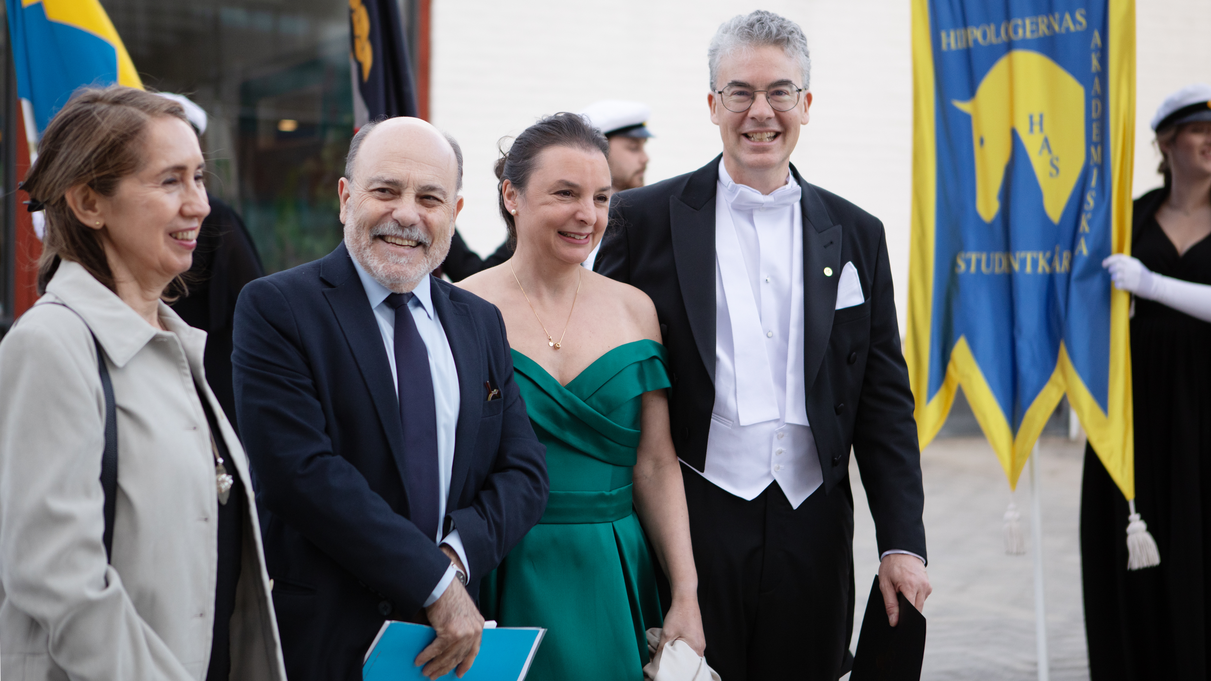 Fyra glada högtidsklädda personer står utomhus omgivna av fanor. 