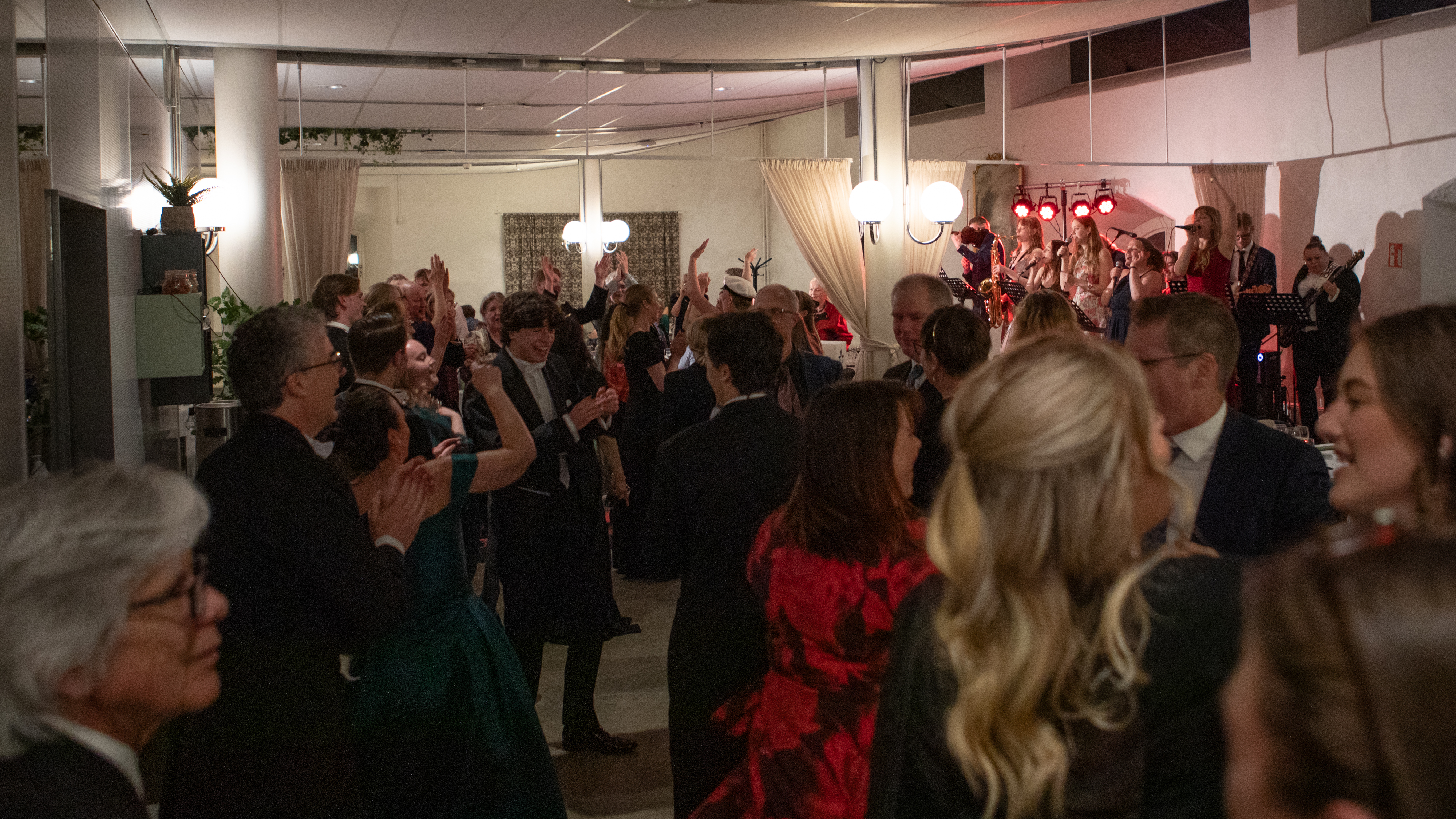 Ett band underhåller på en scen och en publik dansar. Bredvid långa middagsbord med en del sittande gäster. 