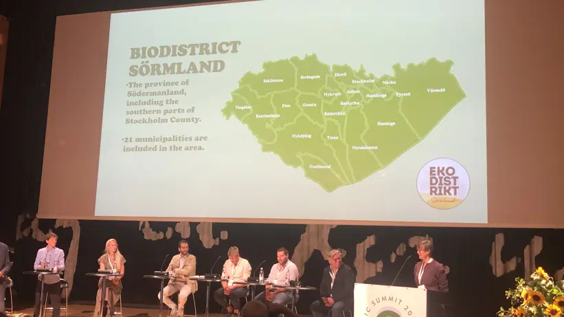 En panel sitter på en stor scen, på en skärm bakom syns rubriken "Ecodistrict Sörmland". Foto.