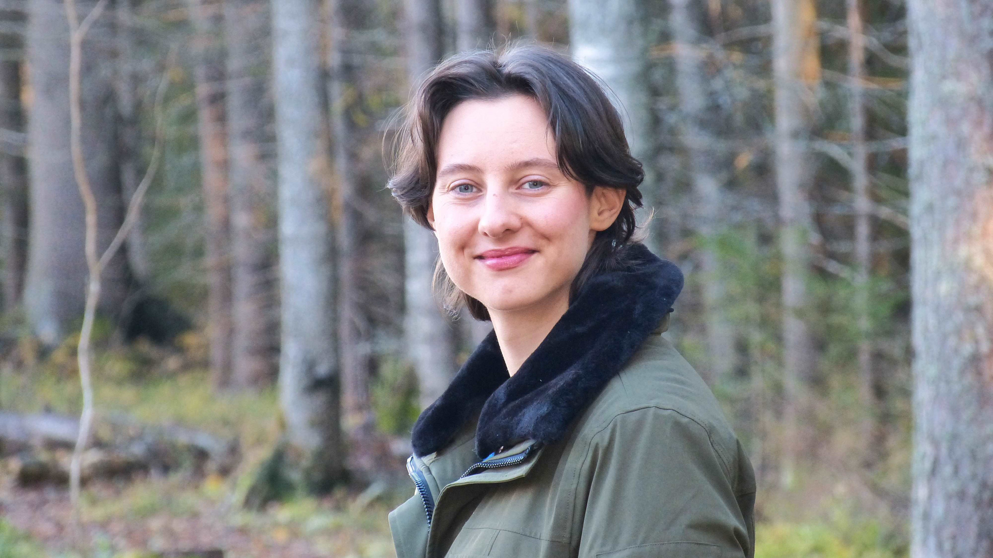 A person with short dark hair and a green jacket stands smiling in a forest with tall trees in the background. The lighting is soft and natural.