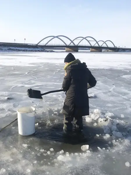 Person stands in an ice hole and pours water into a bucket, with an arch bridge in the background.