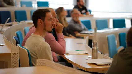 Two men listen attentively in a lecture hall, with more participants blurred in the background. Photo.