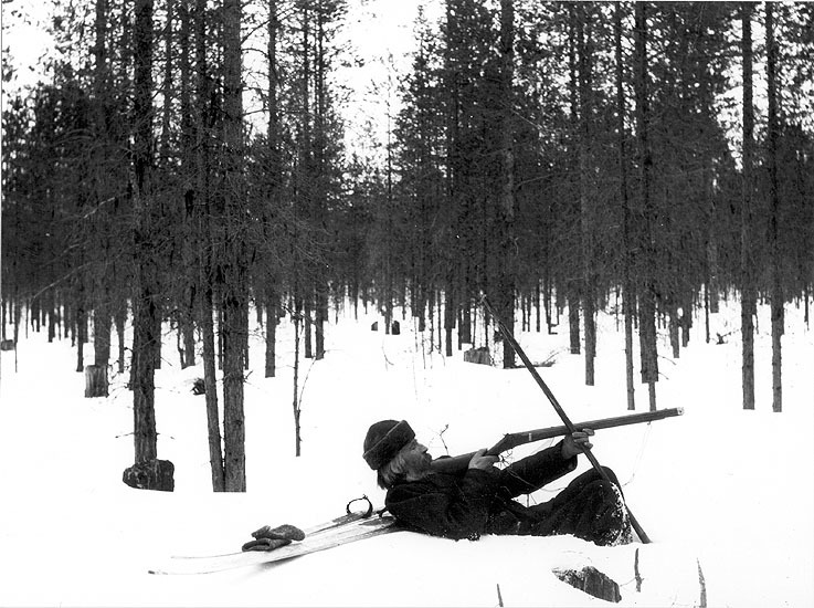En man ligger stödd mot skidorna i ett vinterlandskap med bössan i hand