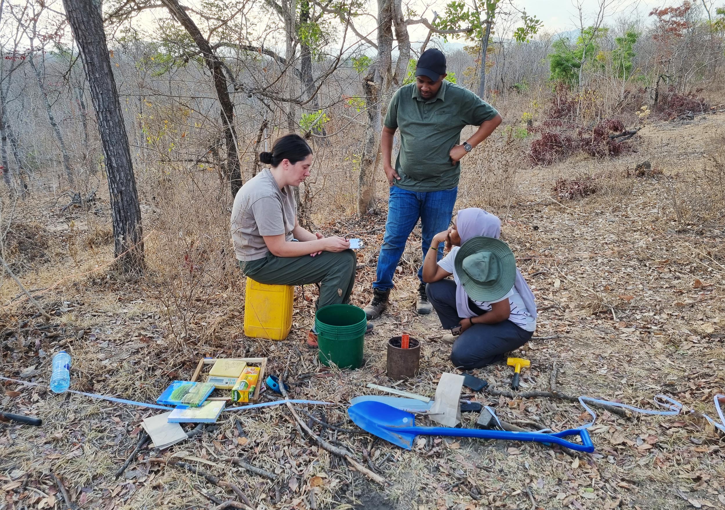 Picture of three persons dressed for fieldwork. They are discussing or pondering over something. One of them, Aida, is sitting on a yellow water can. Another person is squatting next to her and the third person is standing next to them. Around them trees and bushes. On the ground there are spades, buckets and other equipment.  