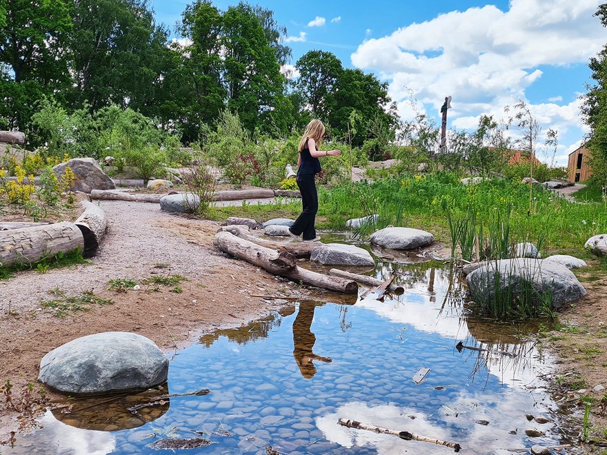 Bild av en flicka som går balansgång på en stock i en park med stockar, små stigar, stenar, planterade ängsblommor och en liten damm. 