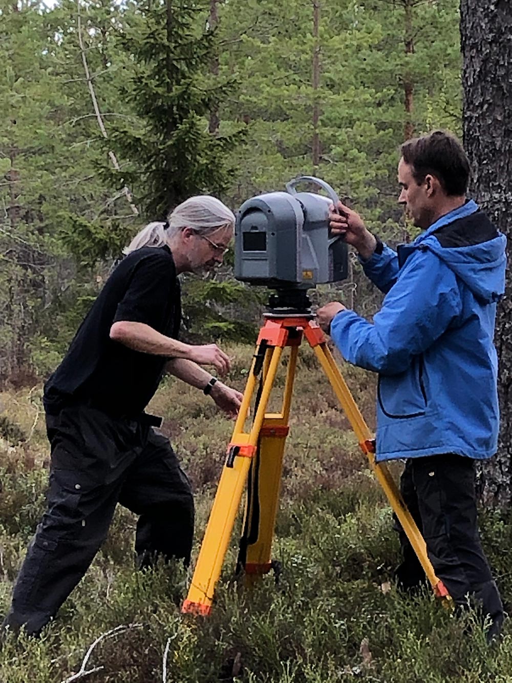 Picture of two persons setting up a grey device with a big yellow tripod in the forest.