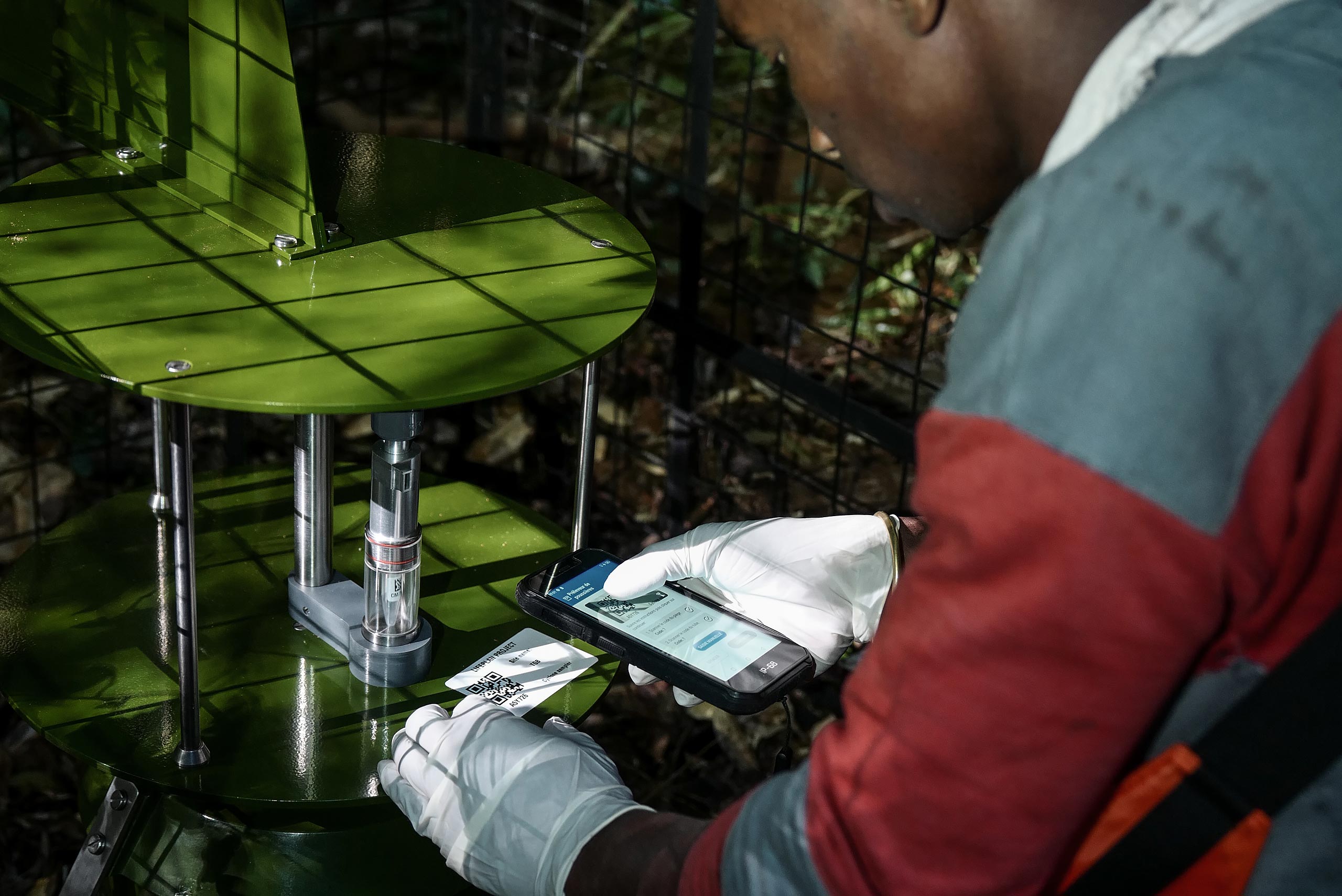 Picture of a man sitting in a forest by a green metal object. The man holds out his phone and scans a code on the object.