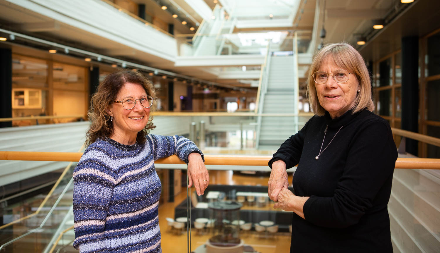 En bild av Karin Wiberg och Agneta Oskarsson som står i ett atrium och tittar in i kameran. I bakgrunden olika våningar, trappor och takfönster i oskärpa.