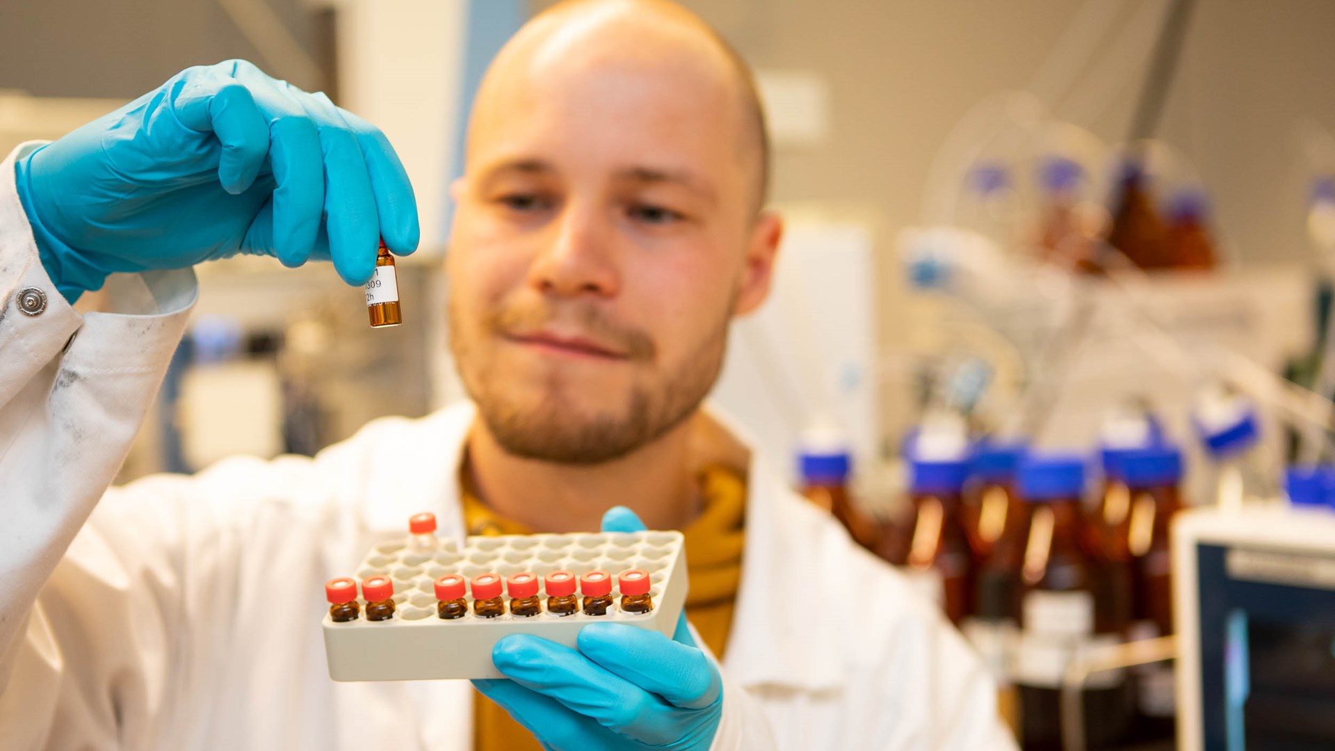 Bild av en person med vit labbrock och blå plasthandskar som håller ett ställ med små provrör i en hand. Med den andra handen lyfter han upp en av de små flaskorna och tittar på den noggrant. I bakgrunden skymtar labbutrustning.