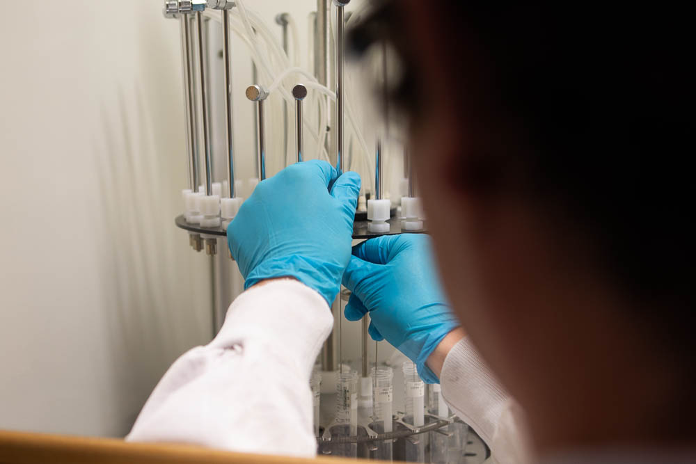 A picture of a person wearing a labcoat and plastic gloves and placing a number of test tubes in a carousel looking device in a fume cupboard. 