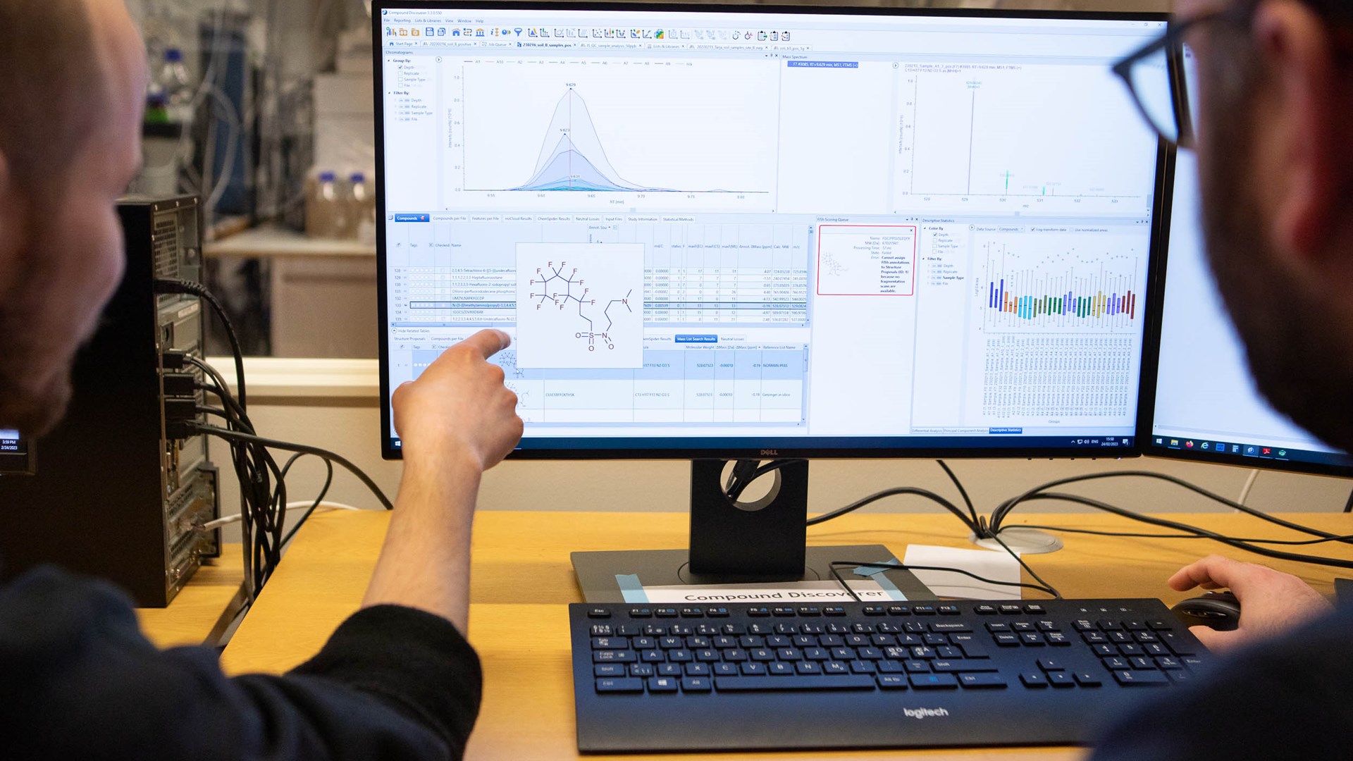 Bild av två personer som sitter vid ett skrivbord och tittar och pekar på en skärm. På skärmen syns ett diagram med en kurva, massor av siffror och en figur som visar den kemiska sammansättningen av något.