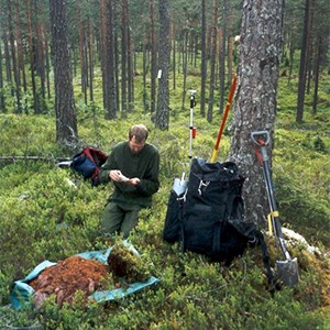 En man sitter i skogen med många verktyg omkring sig, foto.