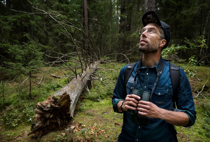 Bild av Marcus Hedblom i skogen med en kikare om halsen.