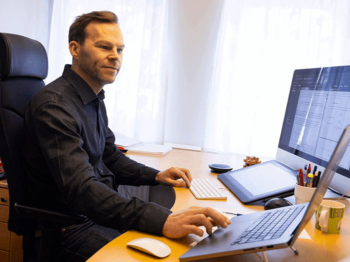 A man in a dark shirt sits in a bright lit office in front of a computer, two screens and a wacom board.