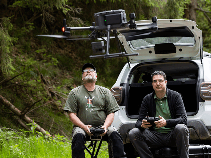 Two persons sitting in open trunk of a car, holding remote controls, one of the persons are looking at a drone hovering in the air.