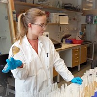 En kvinna med blont hår i hästsvans står i ett laboratorium med en flaska i handen. Foto.