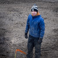 En man i blå jacka står på ett lerigt fält. Foto.