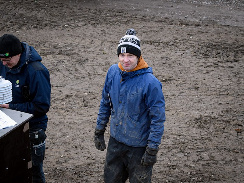 En man i blå jacka står på ett lerigt fält. Foto.