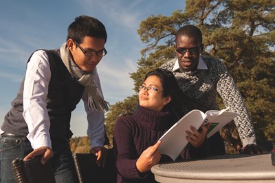 A picture of three people outisde. One of them is sitting down, holding a compendium. The other two is looking at it, all smiling.