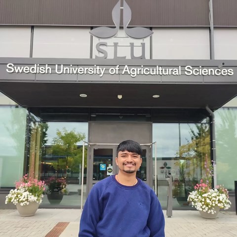 An image of a man standing in front of a house. The house has a sign that says "SLU Swedish University of Agricultural Sciences