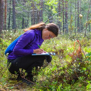 Bilden visar en student som sitter på huk i skogen och skriver på ett papper. 