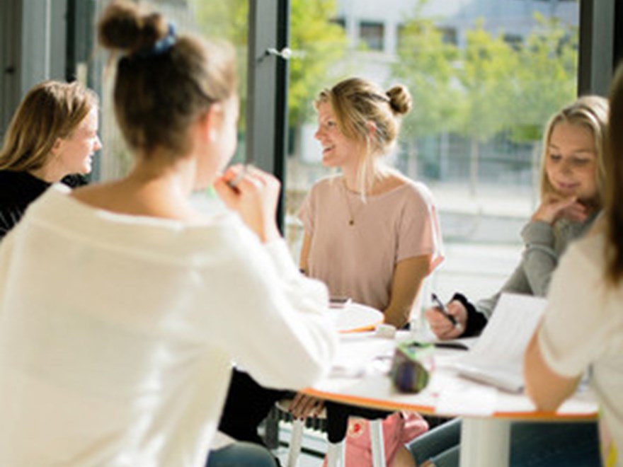 Fem glada studenter runt ett bord framför ett fönster. Foto.