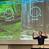 Adam Felton gestikulerar engagerat framför en bild på skog där det står Intensty