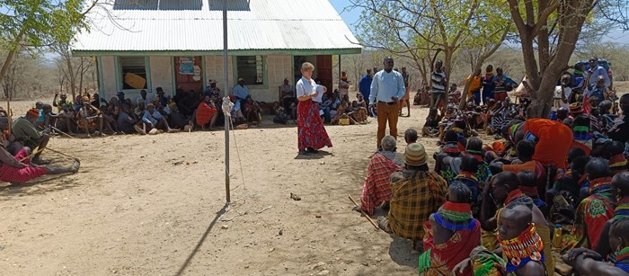 European researcher speaking to men and women gathered under a tree in Uganda.