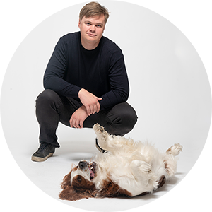 En man sitter på huk bakom en hund som ligger på rygg på golvet framför.
