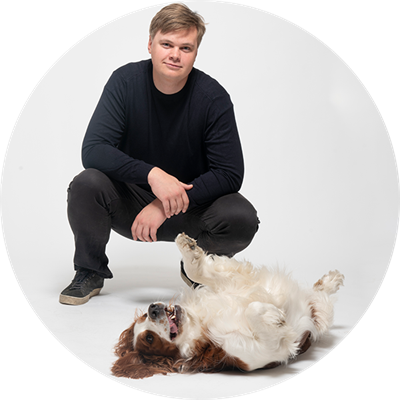 En man sitter på huk bakom en hund som ligger på rygg på golvet framför.