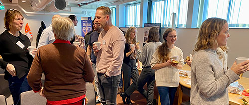 Ett 20-tal personer står och pratar med varandra i ett mötesrum. Foto.
