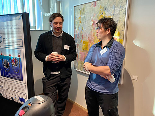 Två unga män står och pratar med varandra, intill en vetenskaplig poster. Foto.