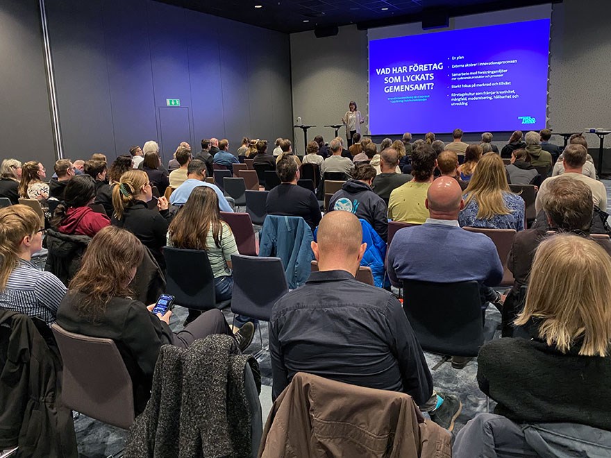 80-tal människor syns bakifrån i en stor konferenssal, med en talare samt en skärm med en presenation på syns längst fram. Foto.