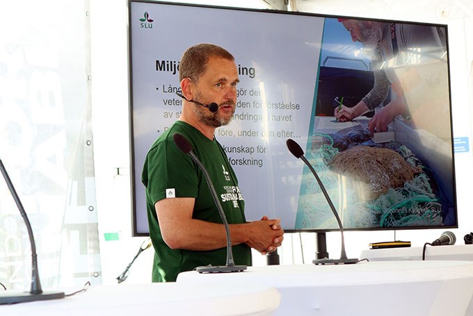 Manlig SLU-forskare med grön SLU T-shirt som står på en scen och håller ett föredrag . I bakgrunden syns en stor skärm som visar powerpointbilder. Foto.
