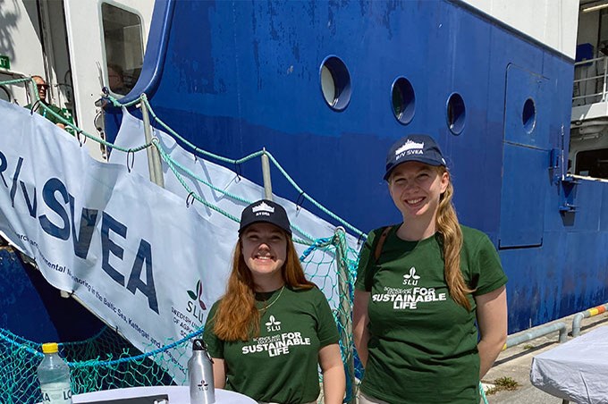 Två unga kvinnor med gröna SLU T-shirts står på en kaj framför ett forskningsfartyg. Foto.