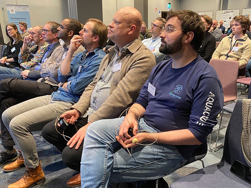 About 80 people sitting in a large conference hall listening to a lecture. Photo.
