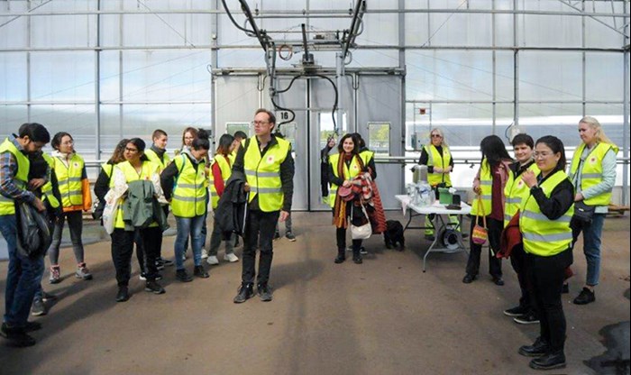 About 20 people are standing in a greenhouse with yellow reflective vests on. Photo.