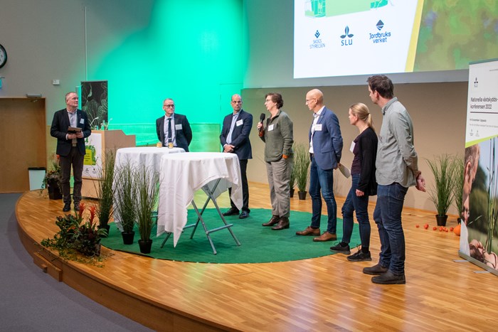 Sju personer står på en scen, en kvinna håller i en mikrofon. Foto.