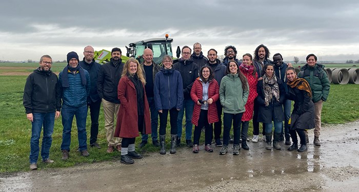 Group photo of about 20 people in a field with a tractor in the background. Photo.