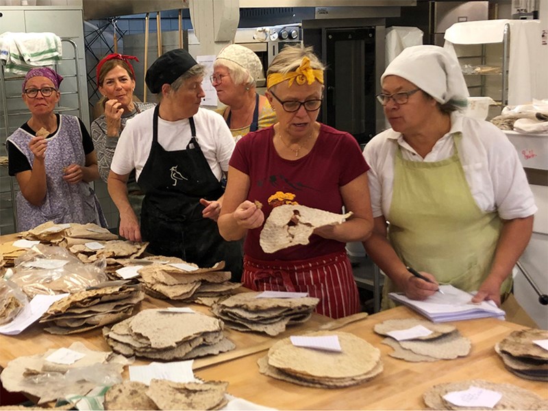 Sju bagare står runt ett bord med tunnbröd, smakar och antecknar i protokoll. Foto.