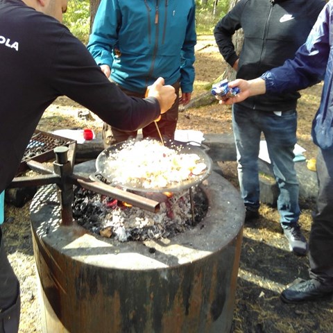 Närbild på en uppbyggd grillplats, med människor som står tätt intill, lagar mat över öppen eld. Foto. 