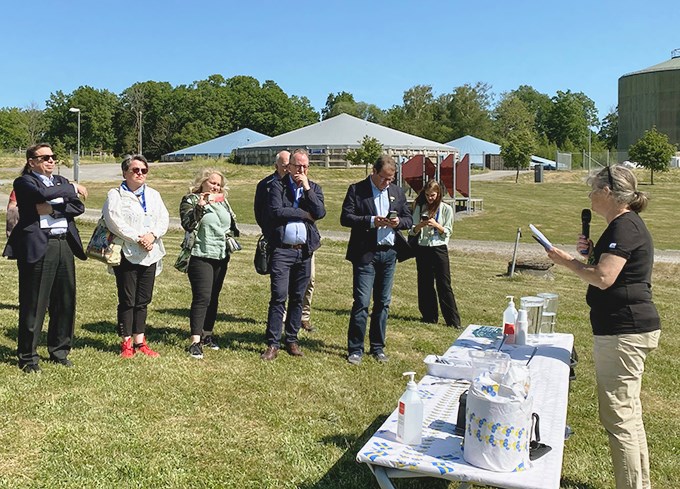 En kvinna pratar i mikrofon för en lyssnande åskådarskara ute på ett fält. Foto. 