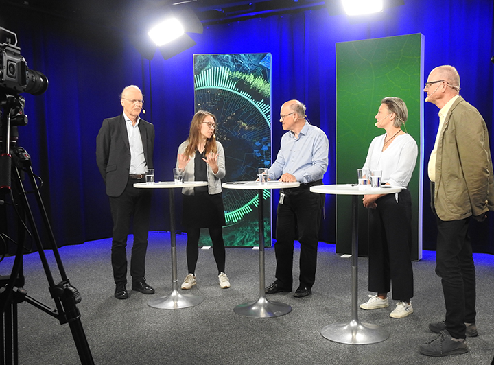 Fem personer står vid bord i studio, en kamera anas i förgrunden. Foto.