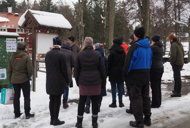 En grupp människor står framför en skylt. En person står vid skylten och berättar vad som beskrivs på den.